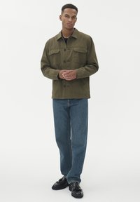 Olivezeleni suknjič s gumbom, z dvema sprednjima žepoma in ovratnikom, v kombinaciji z modrimi jeansi in črnimi vezalkami.