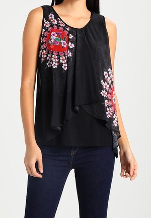 Ärmelloses schwarzes Oberteil mit geschichtetem, asymmetrischem Saum und Blumenmustern mit rosa und weißen Blüten rund um rote Akzente, kombiniert mit dunklen Jeans.