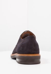 Chaussure en daim bleu marine foncé avec un bout arrondi, des détails cousus et une bordure en cuir brun contrastant autour du col et de la semelle. Semelle en caoutchouc.