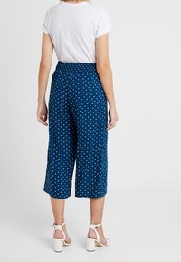 Granatowe kuloty w białe kropki, wykonane z lekkiego materiału, z elastycznym pasem i szerokim krojem nogawek, zestawione z białymi sandałami.