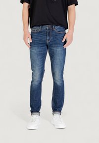 Persona vistiendo jeans azules desgastados con dobladillos remangados y zapatillas blancas, de pie contra un fondo claro.