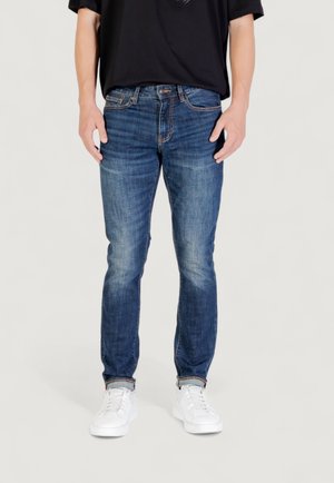Persona che indossa jeans blu sbiaditi con orli risvoltati e sneaker bianche, in piedi contro uno sfondo light uniforme.