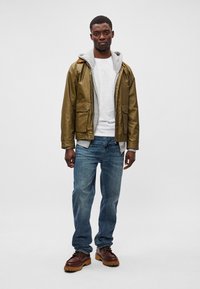 Ελιάς νερού αδιάβροχο μπουφάν με γκρι κουκούλα, λευκό μπλουζάκι, μπλε τζιν και καφέ παπούτσια με κορδόνια και χοντρές σόλες.