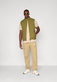Olivgrüne wattierte Weste über einem beige T-Shirt, kombiniert mit hellbraunen Cargo-Hosen. Weiße Sneakers runden das Outfit ab. Heller Hintergrund.