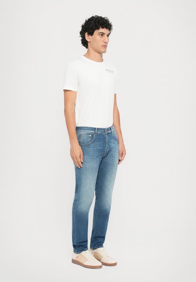 Λεπτά τζιν σε μπλε απόχρωση με ελαφριά φθορά, συνδυασμένα με λευκό βαμβακερό t-shirt και μπεζ αθλητικά παπούτσια με κόκκινες λεπτομέρειες.