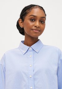 Šviesiai mėlynas marškinėlių mygtukais marškinis su apykakle, pasižymintis lygiu tekstūru ir subtiliu blizgesiu. Priekyje yra maži balti mygtukai.