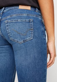 Jeans de mezclilla azul oscuro con un diseño ajustado, presentando un parche de cuero de la marca en la parte trasera de la cintura y un sutil detalle de costura en los bolsillos.