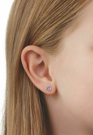 Nahaufnahme eines Ohrs einer Person mit glattem blondem Haar, die einen kleinen silbernen herzförmigen Ohrring mit rosa Steinen trägt.