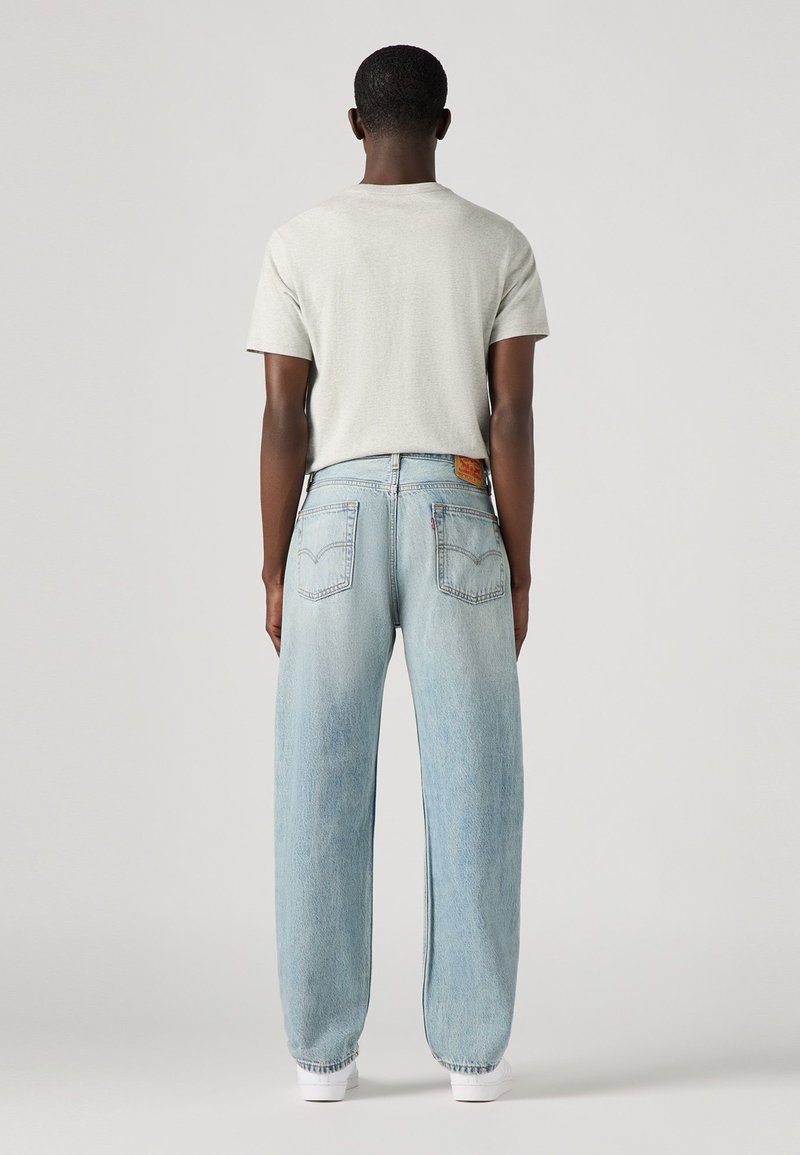 T-shirt gris clair et jeans baggy bleu clair, avec une patch en cuir à l'arrière, accompagnés de baskets blanches. Tissu doux, coupe décontractée.