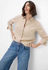 Beige linen button-up shirt with a pocket, three-quarter sleeves, paired with wide-leg blue denim jeans. Simple, casual design.