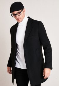 Black wool coat with notched collar, single button, and flap pockets, worn over a white turtleneck. Model wears a black cap and glasses.
