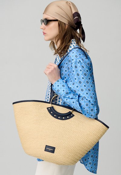 Ampia borsa in paglia a forma rotonda, con dettagli in pelle blu e manici circolari in metallo; il modello indossa una camicia a fantasia blu.
