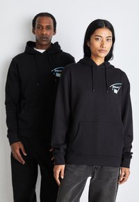 Sudaderas negras con bolsillos frontales, cordones y gráficos del logo. Tela de ajuste holgado y textura suave. Ambos modelos llevan pantalones oscuros.