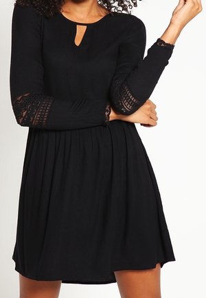 Femme portant une robe noire avec des manches longues en dentelle, un décolleté en forme de goutte et une jupe froncée, posant les bras croisés.