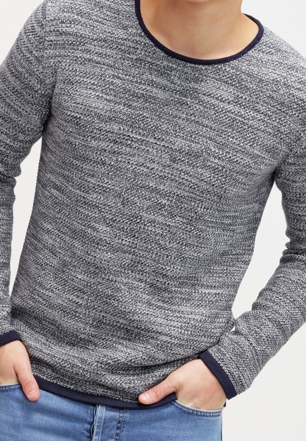 Šedý a námořnicky modrý pletený svetr s texturovaným vzorem, kulatým výstřihem a žebrovanými manžetami. Nosený přes modré džíny, ruce spočívají v kapsách.