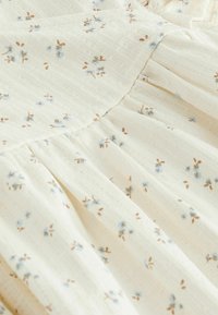 Tissu ivoire clair avec une finition texturée, présentant un imprimé floral éparpillé en bleu et marron. Détails froncés le long des coutures.