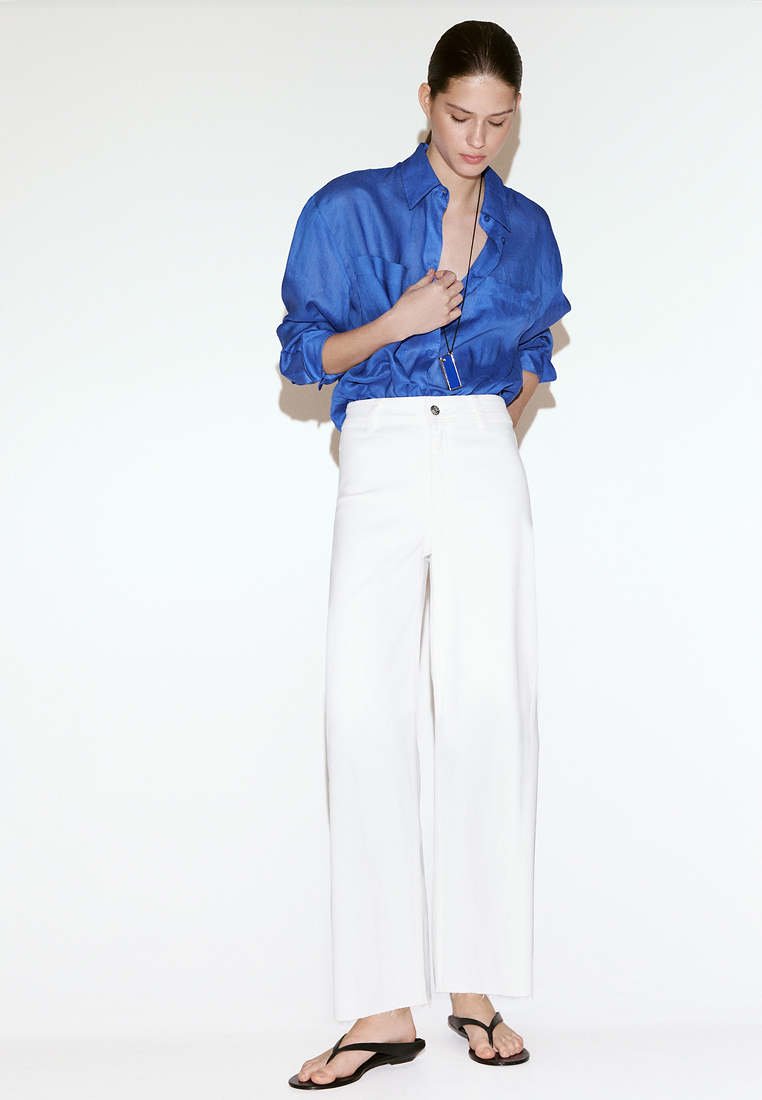 Femme debout portant une chemise bleue à manches longues, un pantalon blanc taille haute à jambes larges, des sandales noires et un long collier.