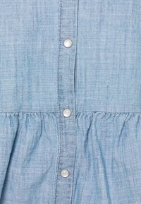 Chemise bleu clair en chambray avec une patte de boutonnage comportant six boutons blancs, une texture froncée à la taille et un tissu tissé doux.