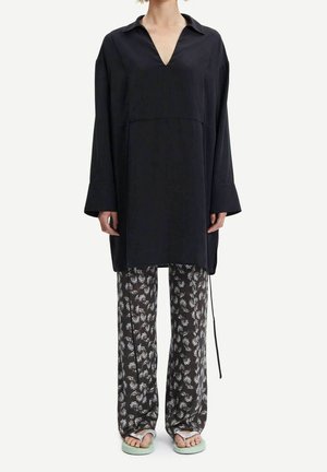 Zwarte tuniek met een V-hals, lange mouwen en een losse fit, gecombineerd met donkergekleurde patroonbroeken met bloemmotieven en lichtgroene sandalen.