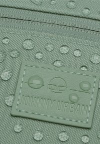 Tessuto impermeabile verde con superficie texture, caratterizzato da punti in rilievo e una toppa con logo cucita. Chiusura con zip nella parte superiore.