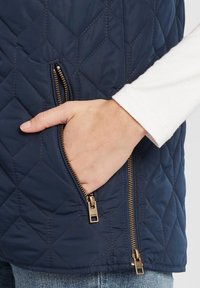 Jūras spēku dabīgā krāsā izšūta veste ar diagonālu rāvējslēdzēja kabatu un zelta detaļām. Audumam ir gluda tekstūra, ko papildina balts krekliņš ar garām piedurknēm.