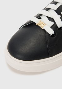 Baskets noires en simili cuir avec des lacets blancs, accents en métal doré, bout arrondi et surface texturée. Semelle en caoutchouc blanche visible.