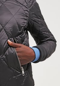 Svart vadderad puffer-jacka med en ficka med dragkedja, ribbade ärmslut och en kontrasterande blå detalj på det inre ärmslutet. Slät yta.