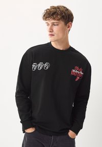 Sort langærmet t-shirt lavet af bomuld, med broderet Rolling Stones læbelogo i hvidt og rødt på brystet, ribstrikkede manchetter.