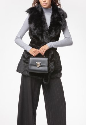 Chaleco de piel sintética negra con un diseño elegante, combinado con un pequeño bolso negro con un cierre en tono dorado. Modelo viste una blusa gris y pantalones a rayas.