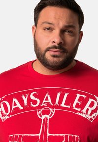 Červené bavlněné tričko s bílým opotřebovaným potiskem slova "DAYSAILER" a grafikou kotvy, design s kulatým výstřihem a krátkými rukávy.