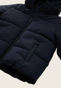 Mornarska napihnjena jakna z zadrgo spredaj, prešita zasnova, ena žep z zadrgo na levem prsnem delu in kapuco. Gladka tekstura in trpežen material.
