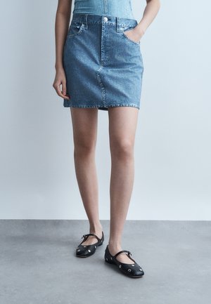 Personne portant une mini-jupe en jean bleu et des ballerines Mary Jane noires avec des œillets argentés, debout avec une main dans la poche sur un sol gris.