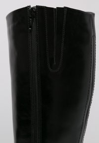 Botte noire en cuir allant jusqu'au genou avec une fermeture éclair sur le côté, présentant une texture lisse et un détail de couture verticale le long des bords.