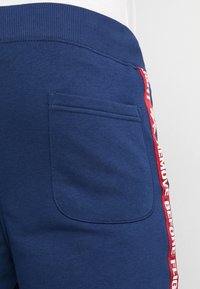 Pantalon de survêtement bleu marine avec une taille côtelée, doté d'une poche latérale et d'une bande rouge avec un texte blanc indiquant "ENLEVEZ AVANT LE VOL."