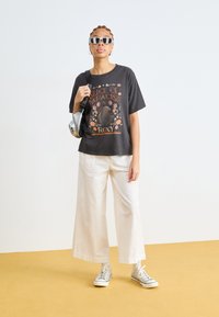 Camiseta gráfica negra con el diseño "Slice of Paradise", combinada con pantalones de pierna ancha blancos y zapatillas plateadas. Incluye gafas de sol y un bolso cruzado.