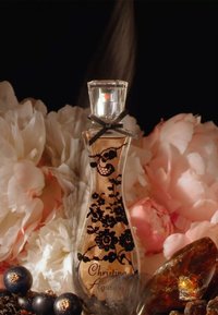 Flacon de parfum élégant avec motif de dentelle noire, entouré de fleurs roses, de cassis et de pierres d’ambre sur un fond sombre.