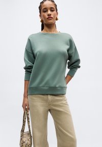 Γυναίκα με πλεξούδες, φοράει πράσινο φούτερ και μπεζ παντελόνι, κρατώντας μια τσάντα με μοτίβο φολιδωτού δέρματος, μπροστά σε ένα απλό φωτεινό φόντο.