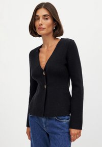 Mustardi V-kaelusega kardigan, mis on valmistatud tekstureeritud kangast, esiosas kahe kuldse nööbiga ja pikkade varrukatega. Kanda siniste teksastega.