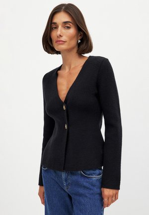 Black V-neck cardigan made of textured fabric, featuring two gold buttons at the front and long sleeves. Worn with blue jeans.
