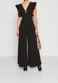 Mujer vestida con un mono negro de pierna ancha con escote en V profundo y mangas con volantes, sosteniendo un bolso beige y usando botas de tacón negras.