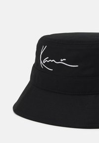 Schwarzes Bucket-Hat aus strukturiertem Stoff mit einem weißen gestickten Logo seitlich. Der Rand ist breit und flach für optimalen Schutz.