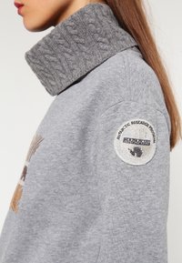 Grijze trui met een dikke kabelgebreide kraag, voorzien van een ronde geborduurde patch op de mouw met een Antarctisch thema en tekst.