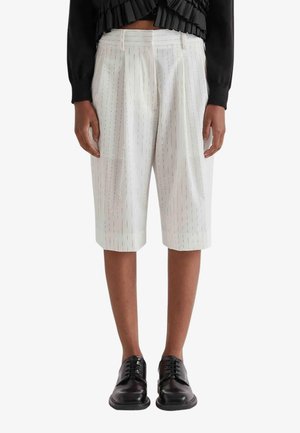 Persona che indossa pantaloncini bianchi a righe fino al ginocchio, top nero a maniche lunghe con volant e scarpe nere con lacci, in piedi dritta.