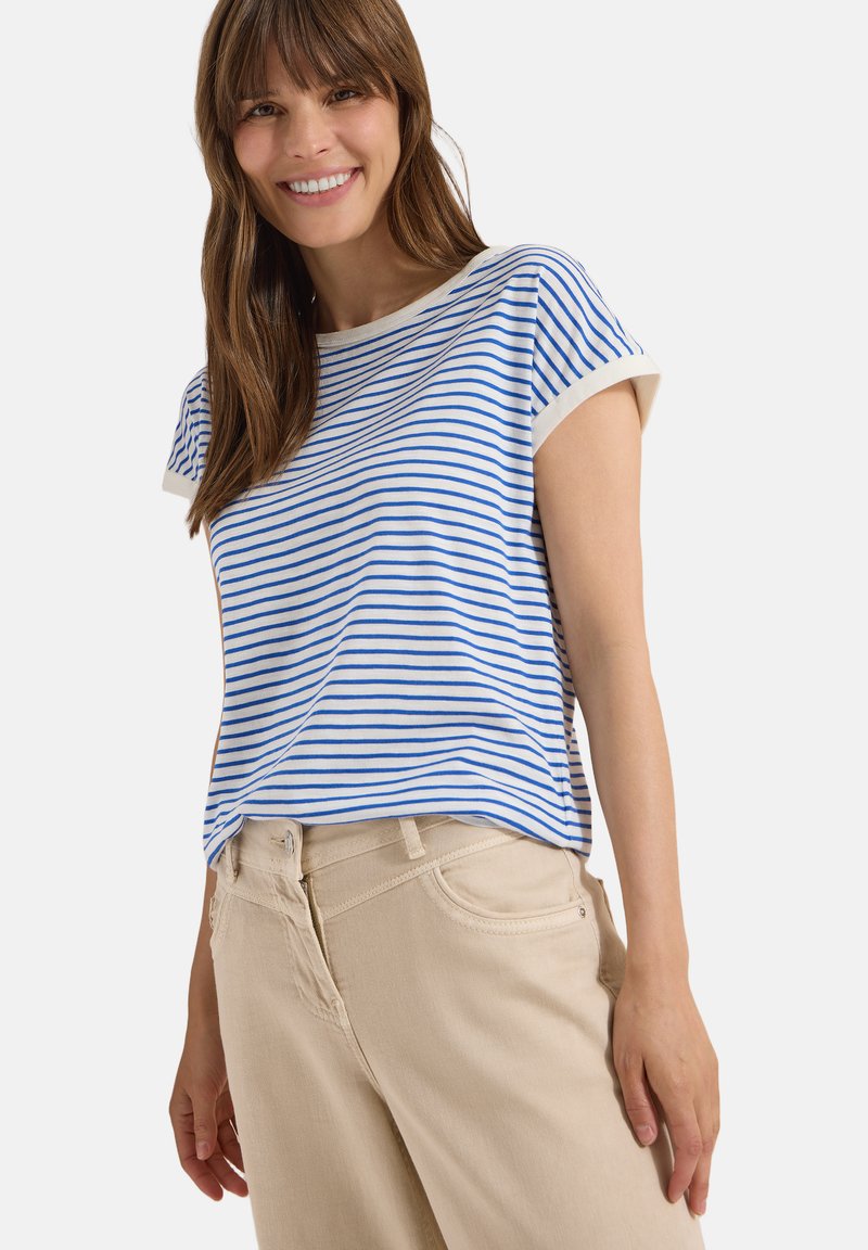 Femme souriante portant un haut blanc à manches courtes avec de fines rayures horizontales bleues et un pantalon beige, debout devant un fond clair uni.