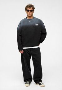 Zwart-grijze gradient sweatshirt met brede mouwen, gelaagd over een wit shirt, gecombineerd met losse zwarte broek en zwarte sneakers.