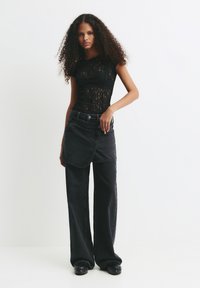 Junge Frau mit lockigem Haar, die ein transparentes schwarzes Spitzentop, einen schwarzen Wickelrock über weit geschnittenen schwarzen Jeans und schwarze Plateauschuhe trägt.