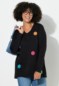 Lachende vrouw met krullend haar draagt een zwarte langemouwen shirt met kleurrijke smiley-patches, blauwe spijkerbroek, zwarte bril en houdt een blauwe handtas vast.