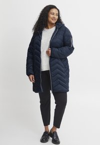 Jūras kara flotes puffer jaka ar šūšanas zīmējumu chevron, ar rāvējslēdzēju un kapuci, kombinēta ar gaiši pelēku džemperi un melniem biksēm. Melni apavi ar sprādzēm.