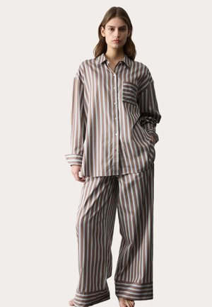 Femme debout pieds nus portant un pyjama ample à rayures verticales marron et blanches avec une poche sur la chemise, sur fond uni.