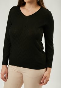 Schwarzer Pullover mit langen Ärmeln und V-Ausschnitt, der ein Zickzackmuster und einen gerippten Saum aufweist. Weiche Textur, kombiniert mit hellbeigen Hosen.